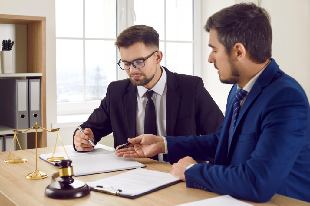 Two men sitting at a table with notebooks, justice scales, and a gavel, going over paperwork, to illustrate the importance of wealth management for lawyers.