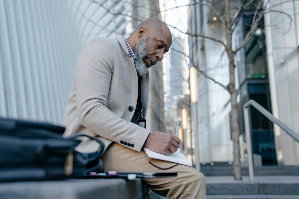 A man sits outside with a notebook and pen, looking thoughtful.