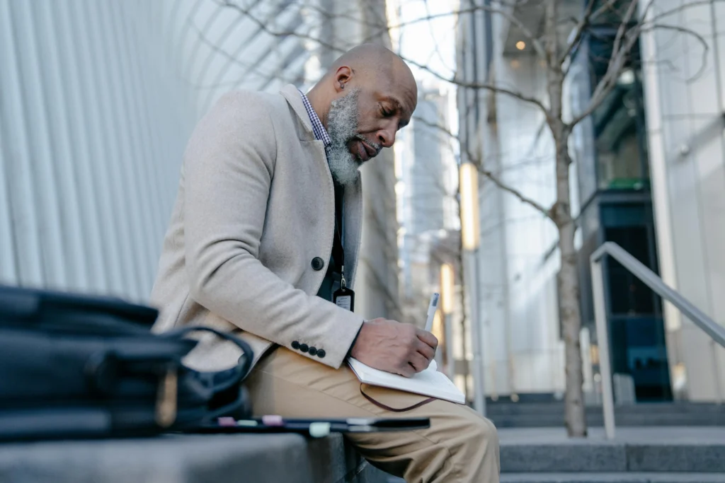 A man sits outside with a notebook and pen, looking thoughtful.